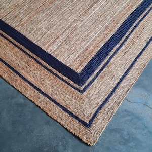 May include: A natural fiber rug with a woven texture and a dark blue border. The rug has a square shape and is made of natural materials.