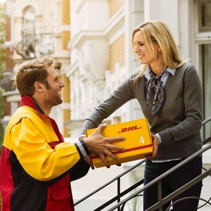 May include: A delivery person in a yellow and red uniform hands a yellow package with the DHL logo to a woman standing on a set of stairs.