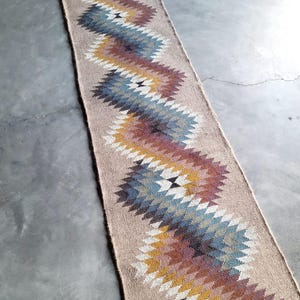 May include: A long, narrow rug with a geometric pattern in shades of brown, blue, and yellow. The rug is woven with a textured, natural fiber.