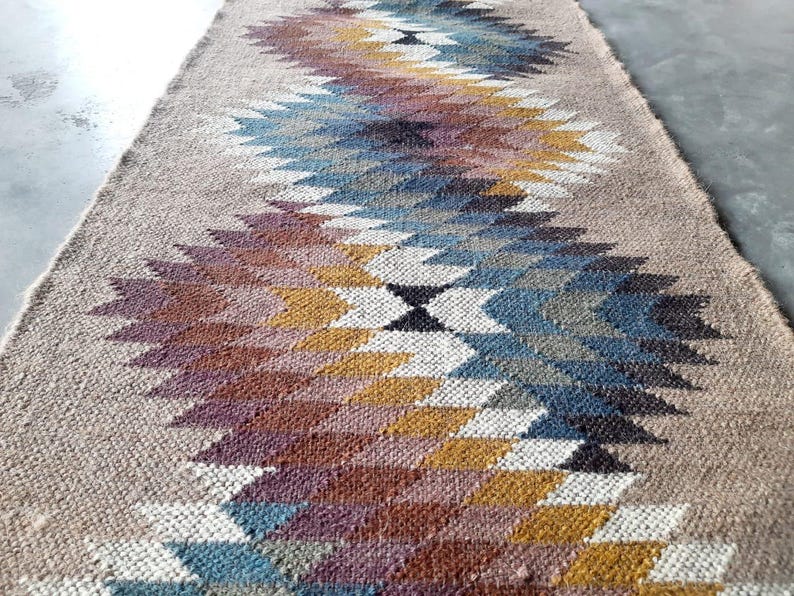 May include: A woven rug with a geometric pattern in shades of brown, blue, white, and gold. The rug has a natural, earthy feel and would add a touch of bohemian style to any room.
