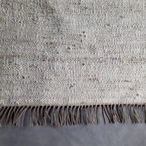 Op de afbeelding: Een close-up van een vloerkleed van natuurlijke vezels met een franje rand. Het vloerkleed is geweven met een textuurpatroon en heeft een licht beige kleur.