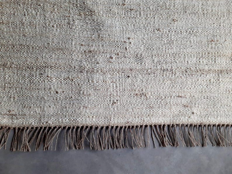 Op de afbeelding: Een close-up van een vloerkleed van natuurlijke vezels met een franje rand. Het vloerkleed is geweven met een textuurpatroon en heeft een licht beige kleur.