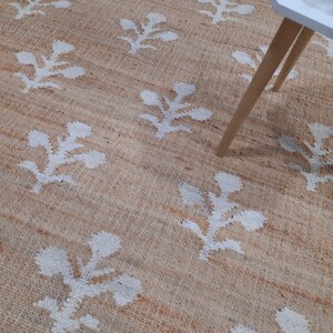 May include: A natural fiber rug with a beige background and a white floral pattern. The rug is shown with a white table with wooden legs.