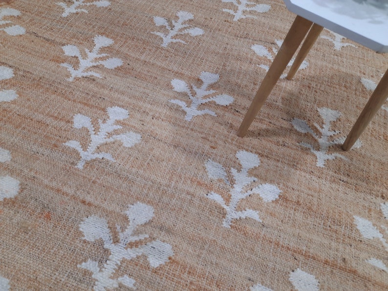 May include: A natural fiber rug with a beige background and a white floral pattern. The rug is shown with a white table with wooden legs.
