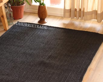 May include: A rectangular black jute rug with visible woven texture. The rug is placed on a light-colored wooden floor. The text "DHURRIETRADE" is printed in white on the rug. The background includes plants, a wooden table, and a window.
