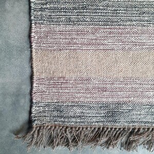 May include: Close-up of the bottom edge of a woven rug with a fringe. The rug has a multi-colored pattern with shades of brown, gray, and pink.