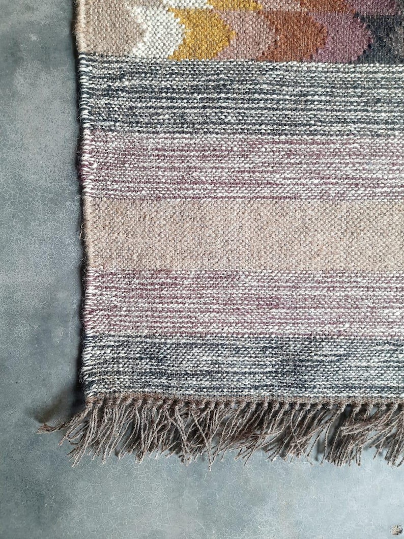 May include: Close-up of the bottom edge of a woven rug with a fringe. The rug has a multi-colored pattern with shades of brown, gray, and pink.