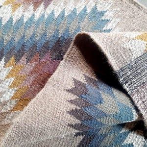 May include: A woven rug with a geometric pattern in shades of brown, blue, and white. The rug is folded in half, showing the intricate details of the design.