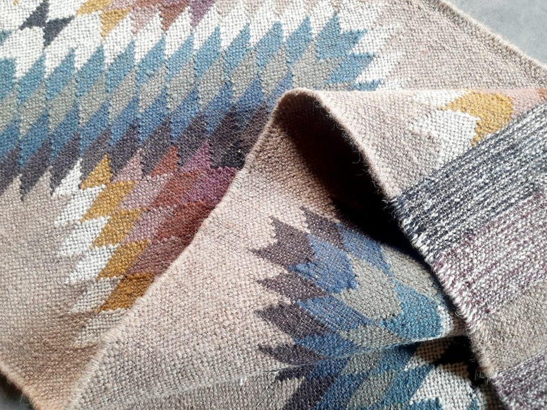 May include: A woven rug with a geometric pattern in shades of brown, blue, and white. The rug is folded in half, showing the intricate details of the design.