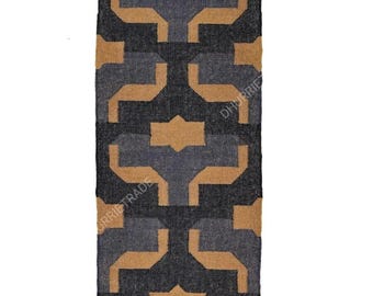 May include: A rectangular rug with a geometric pattern in shades of black, gray, and tan. The rug has a repeating design of interlocking shapes and a fringed edge. The text "DHURRIETRADE" is visible on the rug.