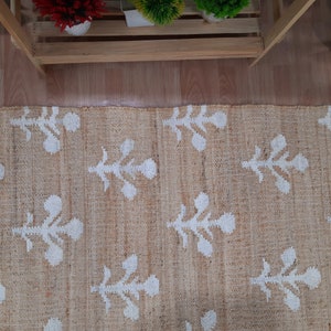 May include: A beige jute rug with a white floral pattern. The rug has a subtle texture and a natural, earthy feel.