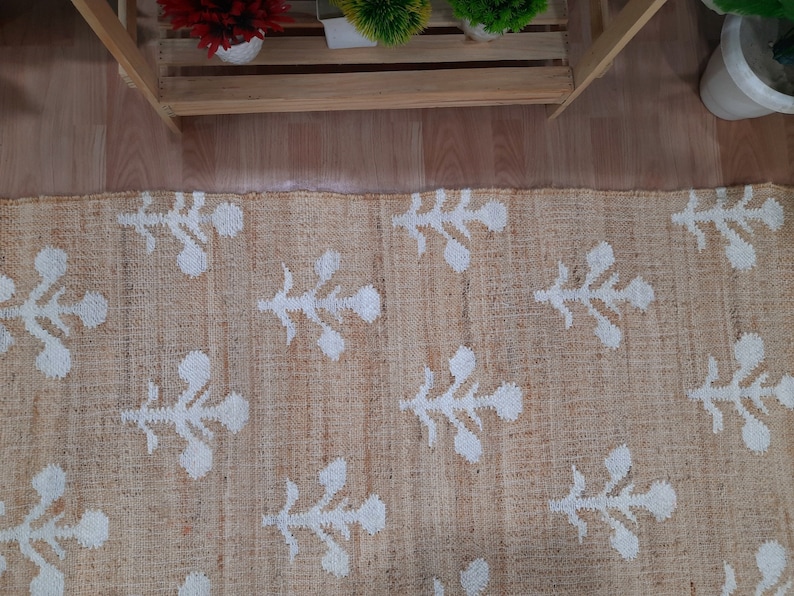 May include: A beige jute rug with a white floral pattern. The rug has a subtle texture and a natural, earthy feel.