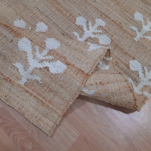 May include: A natural fiber rug with a beige background and white floral patterns. The rug is woven with a textured, slightly rough surface.