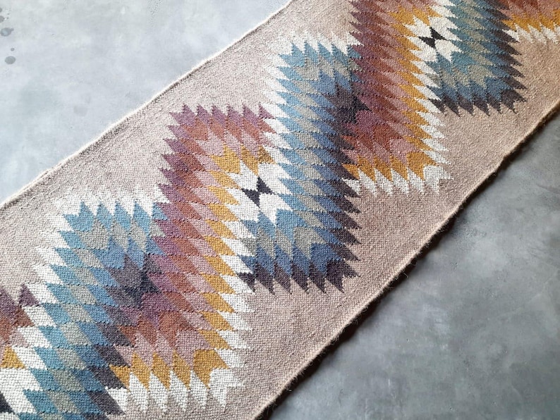 May include: A beige rug with a geometric pattern in shades of blue, brown, and yellow. The pattern is made up of repeating triangles and diamonds.