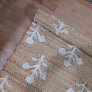 May include: A beige and white woven rug with a floral pattern. The rug is rolled up and the pattern is visible on the rolled up portion.