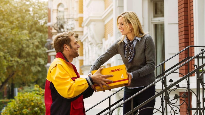 Op de afbeelding: Een bezorger in een geel en rood uniform geeft een geel pakket met het DHL-logo en het nummer 4 aan een vrouw die op een trap staat.