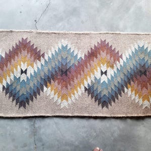 May include: A beige rug with a geometric pattern in shades of brown, blue, and white. The pattern is made up of repeating triangles and diamonds.