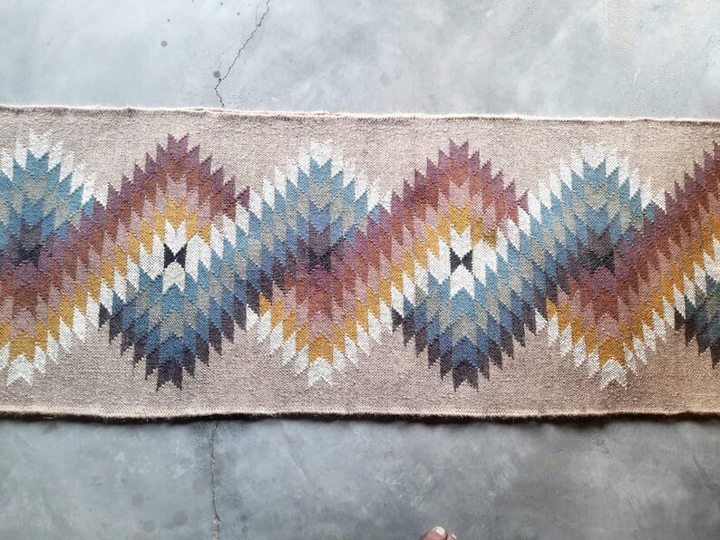 May include: A beige rug with a geometric pattern in shades of brown, blue, and white. The pattern is made up of repeating triangles and diamonds.