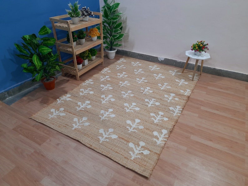 May include: A beige jute rug with a white floral pattern. The rug is on a wooden floor in a room with a wooden shelf and potted plants.