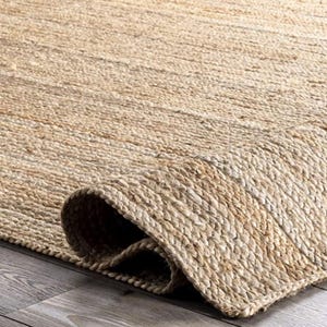 Peut inclure: Un tapis en fibres naturelles avec un bord enroulé. Le tapis est fait de fibres de jute tissées dans des tons de beige et de beige. La texture du tapis est visible, avec une surface légèrement inégale. Le tapis semble être sur un sol en bois.