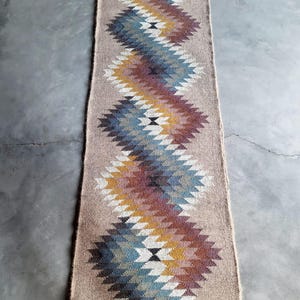 May include: A long, narrow rug with a geometric pattern in shades of blue, brown, white, and yellow. The rug has a woven texture and is made of natural fibers.