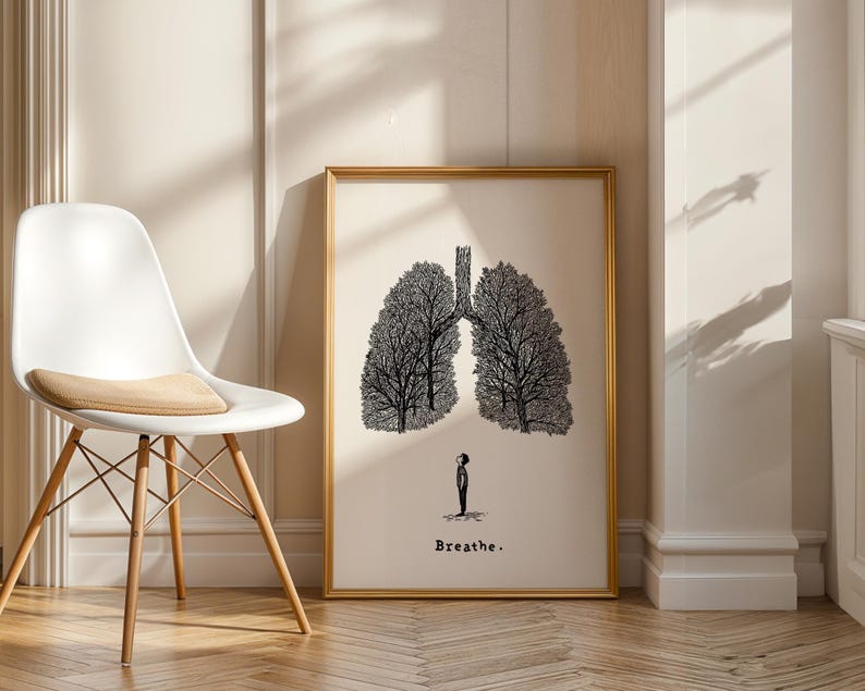 May include: A framed art print featuring a black and white illustration of lungs shaped like trees, with a small figure beneath. The word "Breathe" is printed below the figure. The artwork is displayed in a room with a white chair.
