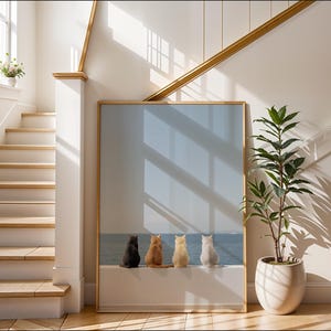 May include: A framed print depicting four cats of varying colours, positioned together, gazing at a blue ocean view. The artwork is displayed in a bright room with a staircase and a potted plant.