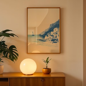 May include: A framed print of a coastal scene with a sailboat and buildings, displayed above a wooden cabinet. A round, glowing lamp and potted plants are on the cabinet. The print has a wooden frame and a blue and white color scheme.