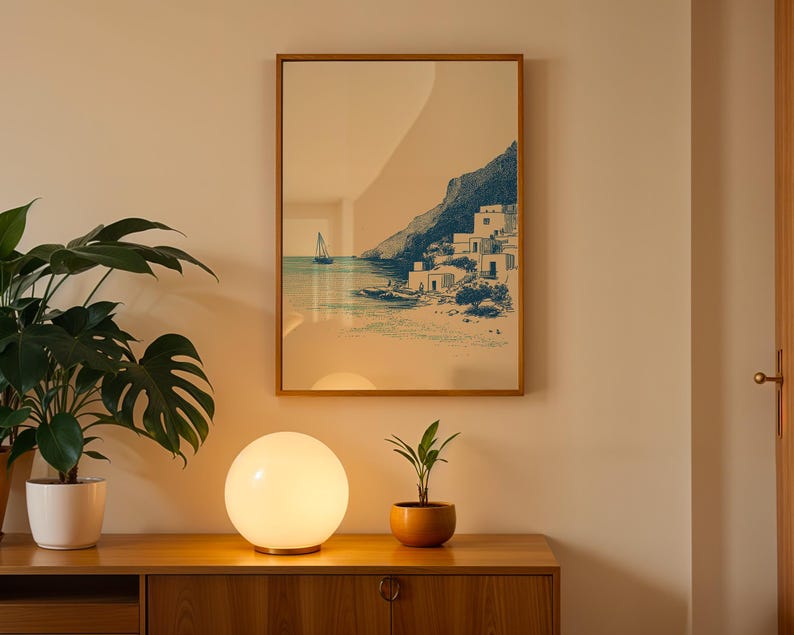 May include: A framed print of a coastal scene with a sailboat and buildings, displayed above a wooden cabinet. A round, glowing lamp and potted plants are on the cabinet. The print has a wooden frame and a blue and white color scheme.