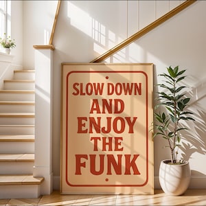May include: A framed sign with the text "SLOW DOWN AND ENJOY THE FUNK" in red letters. The sign is beige with a red border and is leaning against a wall. A potted plant sits to the right.