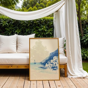 May include: A framed art print depicting a coastal scene with a sailboat, white buildings, and a mountain. The artwork is in shades of blue and beige, with a wooden frame. The print is displayed outdoors on a wooden deck.