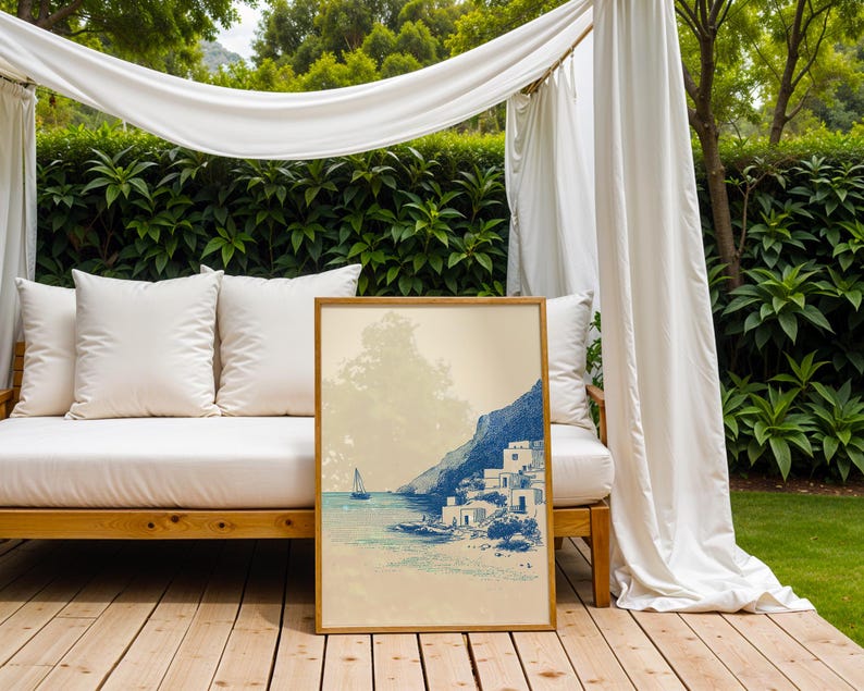 May include: A framed art print depicting a coastal scene with a sailboat, white buildings, and a mountain. The artwork is in shades of blue and beige, with a wooden frame. The print is displayed outdoors on a wooden deck.