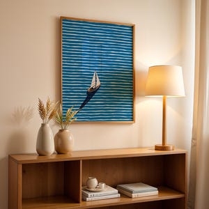 Puede incluir: Una obra de arte enmarcada representa un velero sobre un fondo a rayas azules y blancas. La obra de arte cuelga sobre un mueble de madera con una l&aacute;mpara y dos jarrones. La l&aacute;mpara tiene una pantalla beige y una base de madera. Los jarrones son beige y blancos.