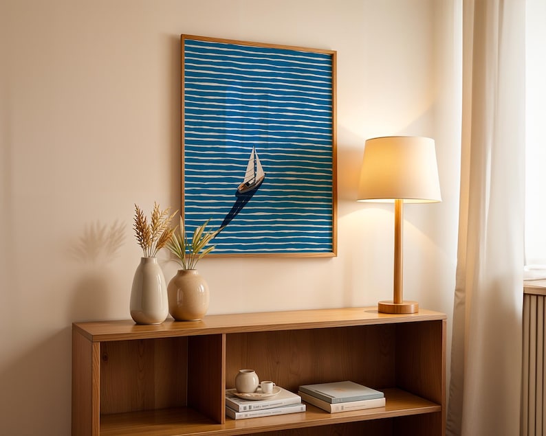 Puede incluir: Una obra de arte enmarcada representa un velero sobre un fondo a rayas azules y blancas. La obra de arte cuelga sobre un mueble de madera con una l&aacute;mpara y dos jarrones. La l&aacute;mpara tiene una pantalla beige y una base de madera. Los jarrones son beige y blancos.