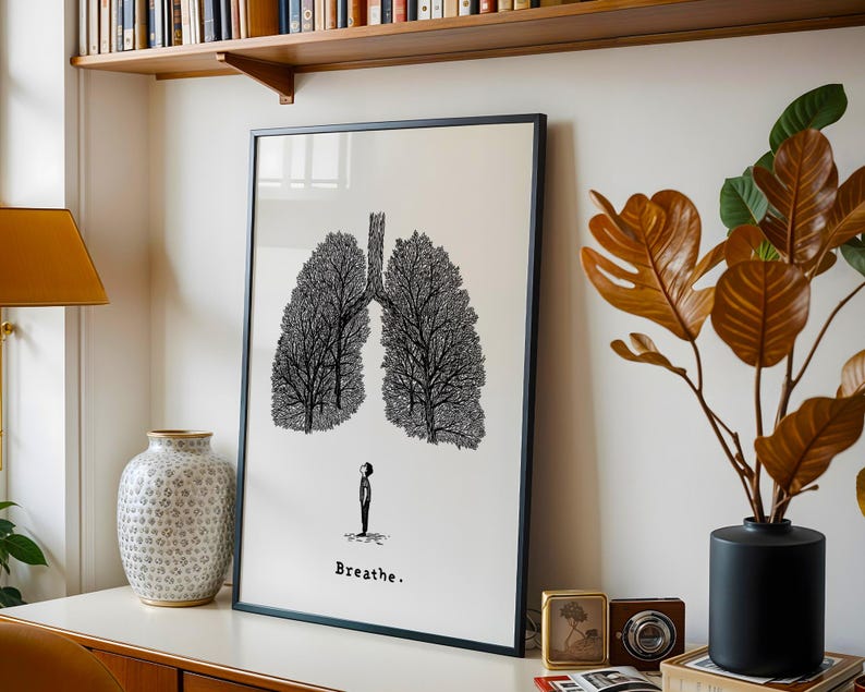 May include: Black framed art print featuring a stylized illustration of lungs formed by trees. A small figure stands below the lungs, with the word "Breathe" printed beneath. The artwork is displayed on a white wall, with a wooden shelf above.