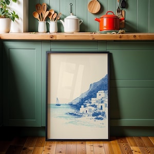 May include: A framed print featuring a coastal scene with a sailboat, white buildings, and a mountain in shades of blue. The artwork is displayed against a backdrop of a green kitchen cabinet and wooden floor.