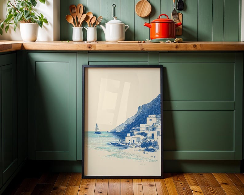 May include: A framed print featuring a coastal scene with a sailboat, white buildings, and a mountain in shades of blue. The artwork is displayed against a backdrop of a green kitchen cabinet and wooden floor.
