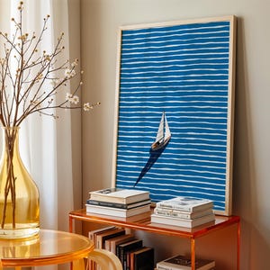Puede incluir: Una impresi&oacute;n art&iacute;stica enmarcada con un velero sobre un fondo a rayas azules y blancas. La obra de arte se exhibe sobre una mesa naranja transparente con libros y un jarr&oacute;n amarillo con ramas.