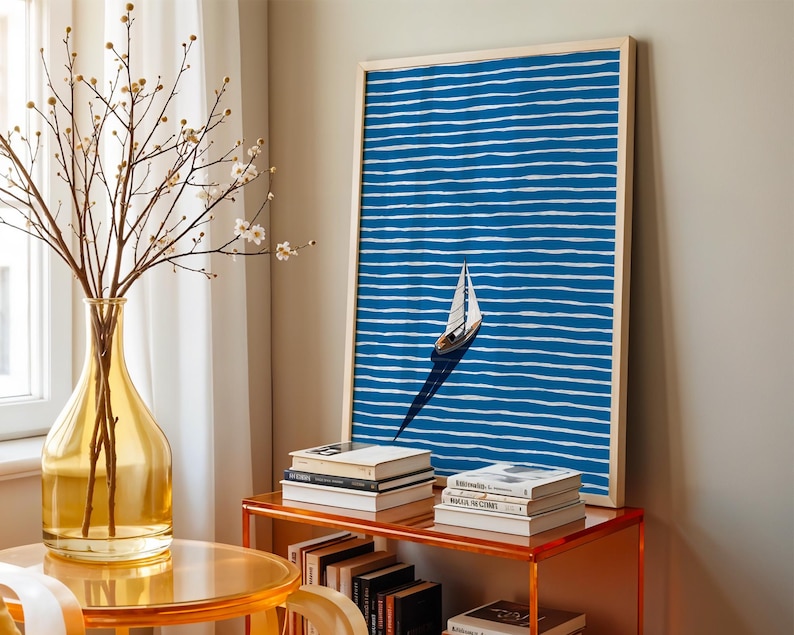 Puede incluir: Una impresi&oacute;n art&iacute;stica enmarcada con un velero sobre un fondo a rayas azules y blancas. La obra de arte se exhibe sobre una mesa naranja transparente con libros y un jarr&oacute;n amarillo con ramas.
