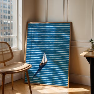 Puede incluir: Una impresi&oacute;n art&iacute;stica enmarcada que presenta un velero sobre un fondo a rayas azules y blancas. La obra de arte est&aacute; apoyada contra una pared. Se ven una silla de madera y una ventana. La impresi&oacute;n tiene un marco de madera.
