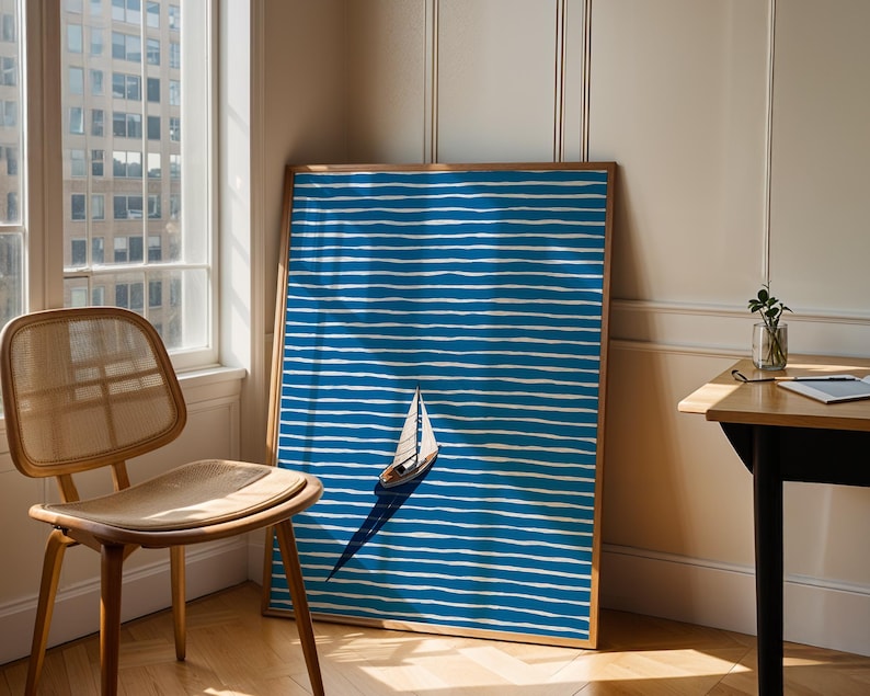 Puede incluir: Una impresi&oacute;n art&iacute;stica enmarcada que presenta un velero sobre un fondo a rayas azules y blancas. La obra de arte est&aacute; apoyada contra una pared. Se ven una silla de madera y una ventana. La impresi&oacute;n tiene un marco de madera.