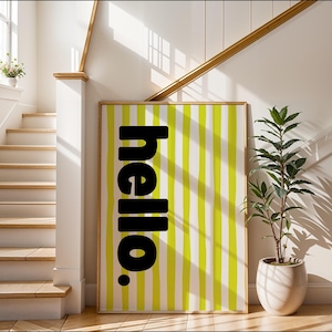 Op de afbeelding: Een ingelijste kunstprint met het woord "hello." in zwart tegen een achtergrond van verticale, handgetekende, gele en witte strepen. Het kunstwerk wordt tentoongesteld in een lichte ruimte met een trap en een plant in een pot.