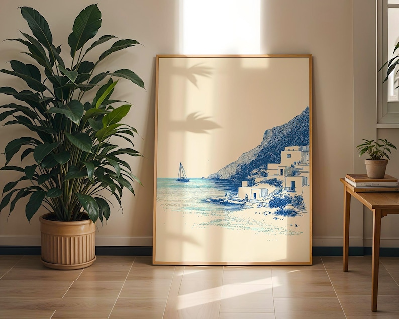 May include: A framed art print depicting a coastal scene with a sailboat on the water and buildings on a hillside. The artwork is in shades of blue and beige, with a wooden frame. A large potted plant is to the left.