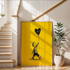 May include: A framed art print with a black and white illustration of a child holding a heart-shaped balloon on a yellow background. The artwork is displayed in a room with stairs and a potted plant.
