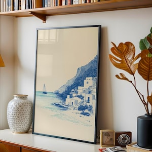 May include: A framed print featuring a coastal scene with a sailboat, buildings, and a mountain in blue and white tones. The artwork is displayed on a white surface, alongside a decorative vase, a plant, and other decor.