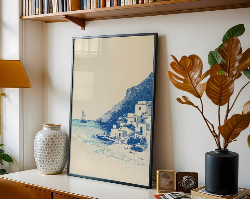 May include: A framed print featuring a coastal scene with a sailboat, buildings, and a mountain in blue and white tones. The artwork is displayed on a white surface, alongside a decorative vase, a plant, and other decor.