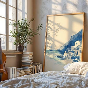 May include: A framed print of a coastal scene with blue and white buildings, a mountain, and a sailboat. The artwork is displayed in a room with a cello, a potted plant, and a stack of books. The room has a large window and a bed with white bedding.