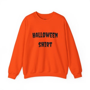Może przedstawiać: Jasnopomarańczowa bluza z napisem "HALLOWEEN SHIRT" wydrukowanym czarnymi, pogrubionymi literami. Top z długim rękawem jest w jednolitym kolorze, idealny na sezon Halloween. Swobodny, wygodny element garderoby.