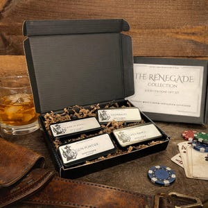 May include: A black gift box containing four solid colognes from "The Renegade Collection." Each cologne tin features a cowboy graphic and names like "Desperado" and "Gun Powder." The box is open, revealing the colognes nestled in brown packing material.