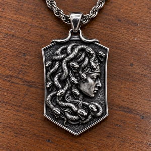 Puede incluir: Un colgante de tono plateado con un relieve detallado de una figura con serpientes por cabello, presentado en un marco con forma de escudo. El colgante cuelga de una cadena de metal grueso y retorcido, mostrando texturas y profundidad.